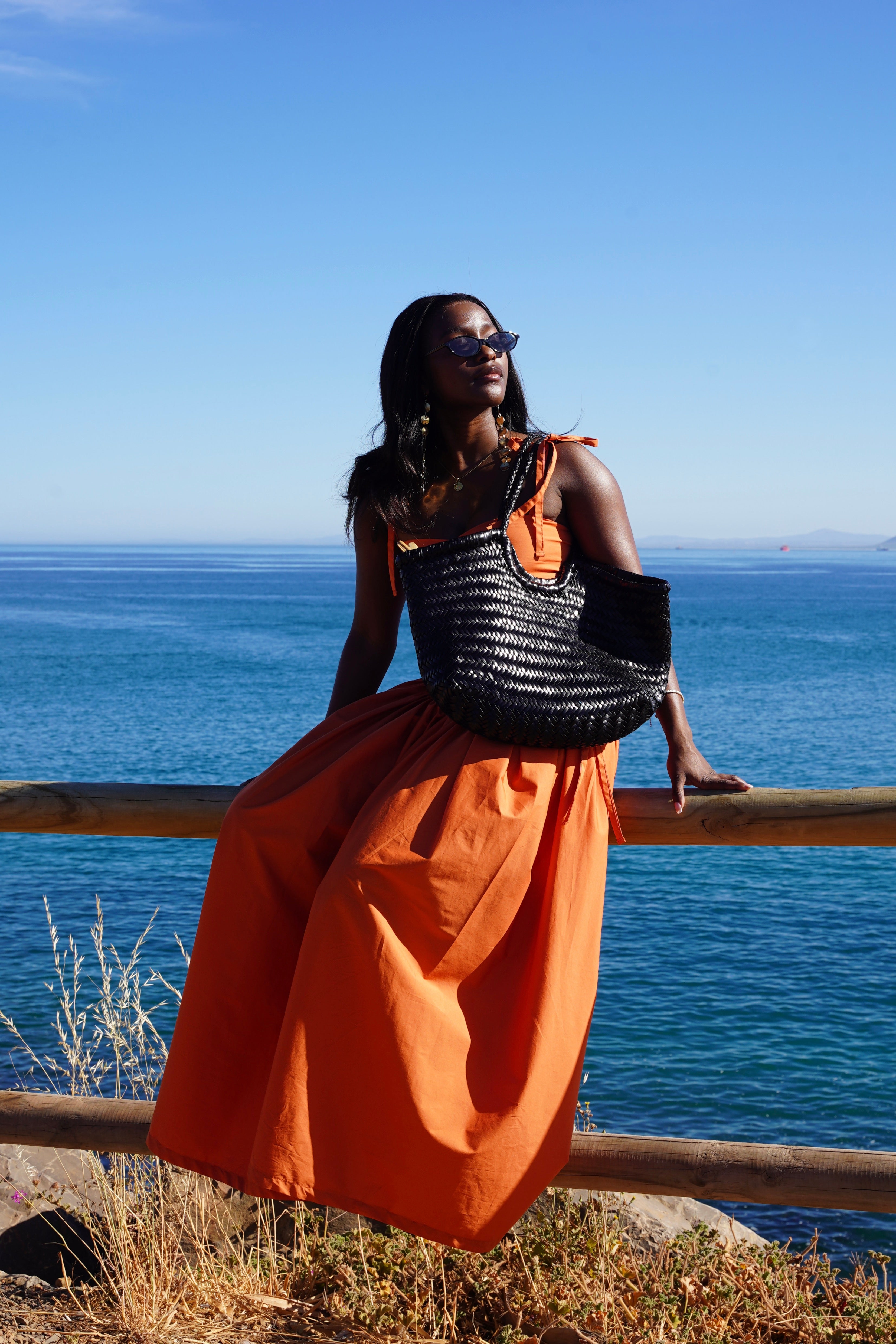 Woman in an orange dress and black top standing by a wooden railing with a blue ocean and sky in the background.
