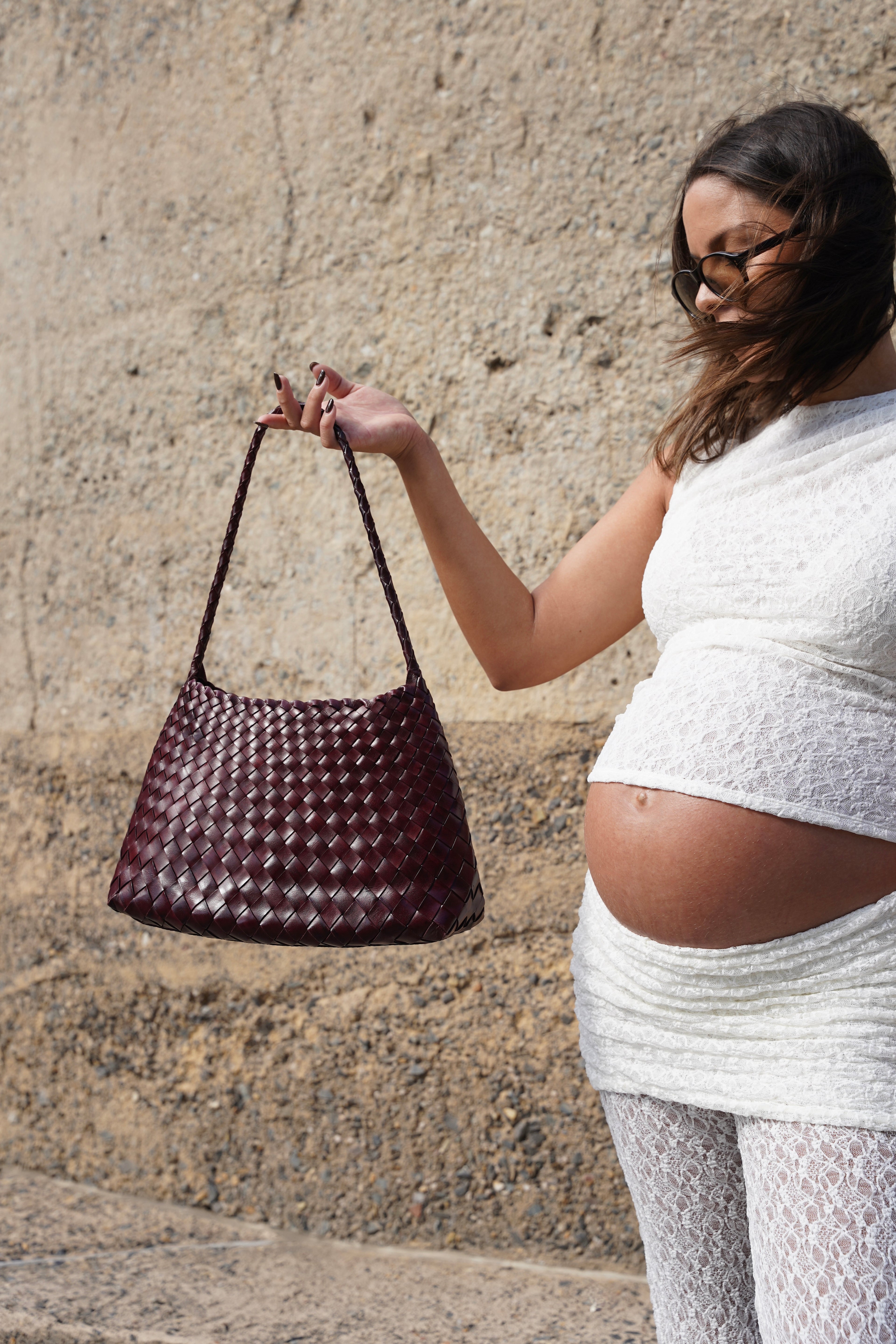 Pregnant woman holding a woven handbag against a stone wall.