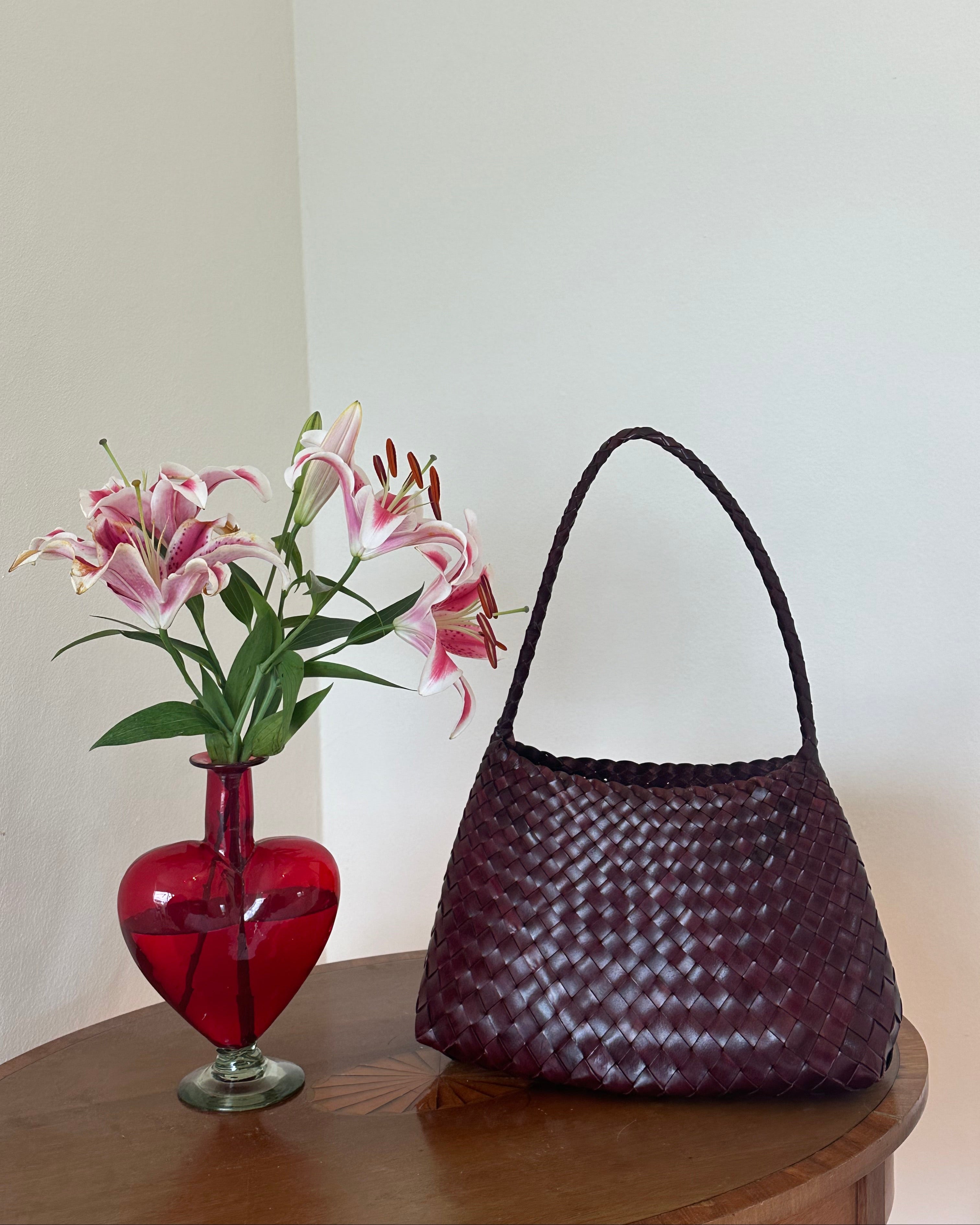 Red glass vase with pink flowers and a woven handbag on a wooden surface.