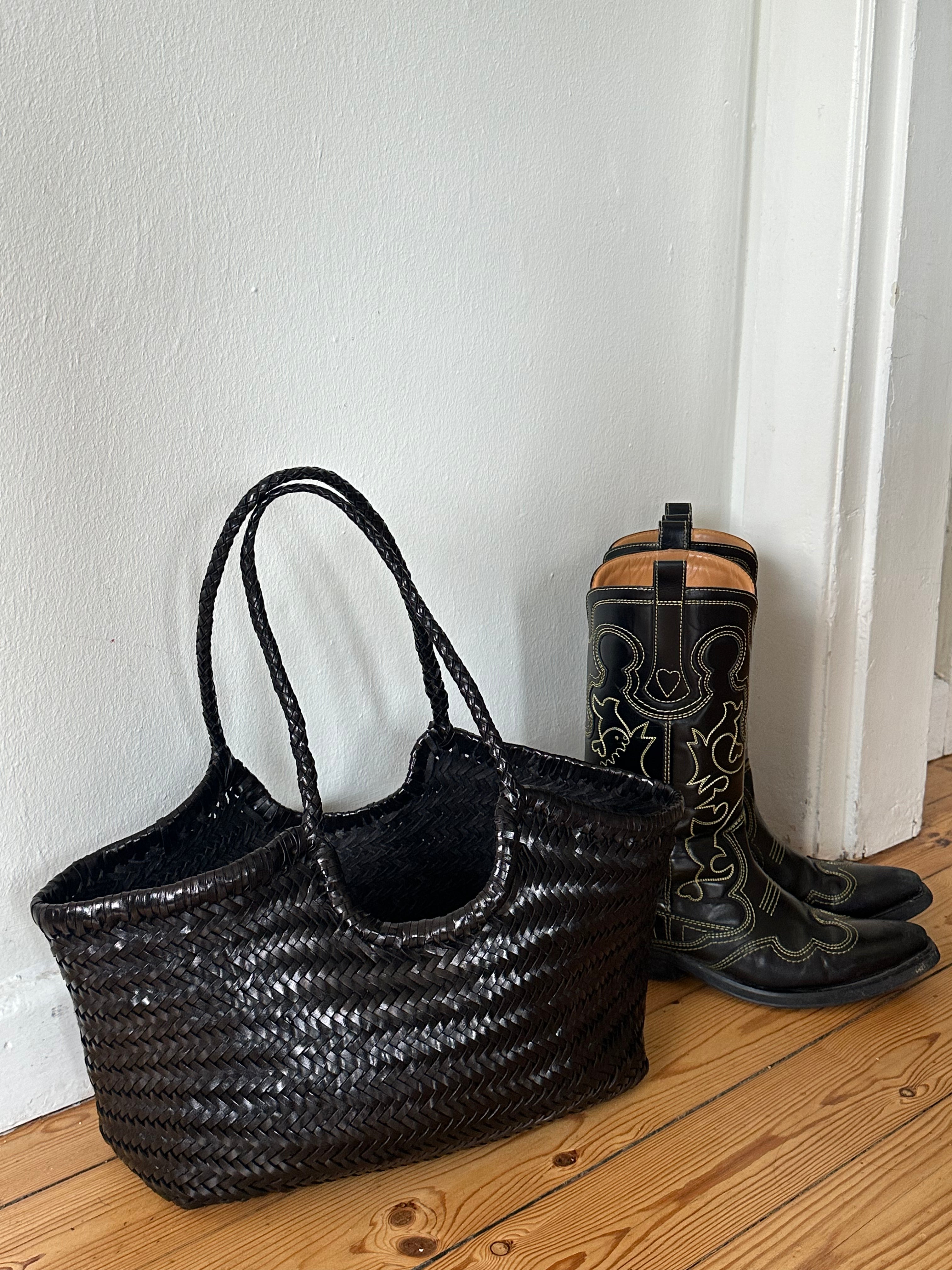 Black woven bag and black boots on a wooden floor against a white wall.