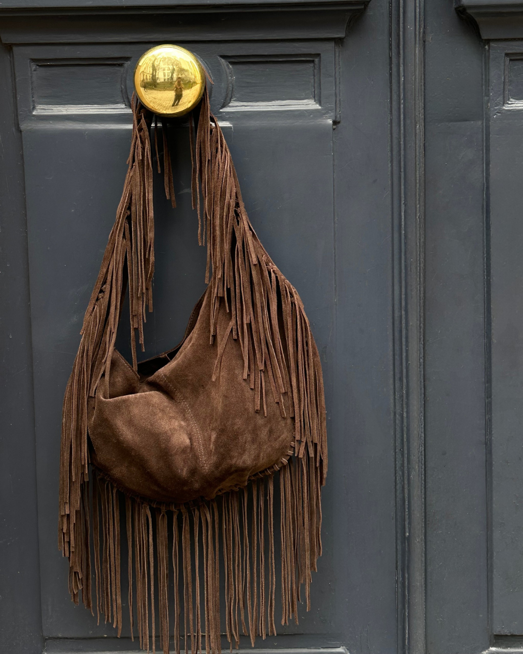 Brown suede handbag with fringes hanging on a dark door with a gold doorknob.